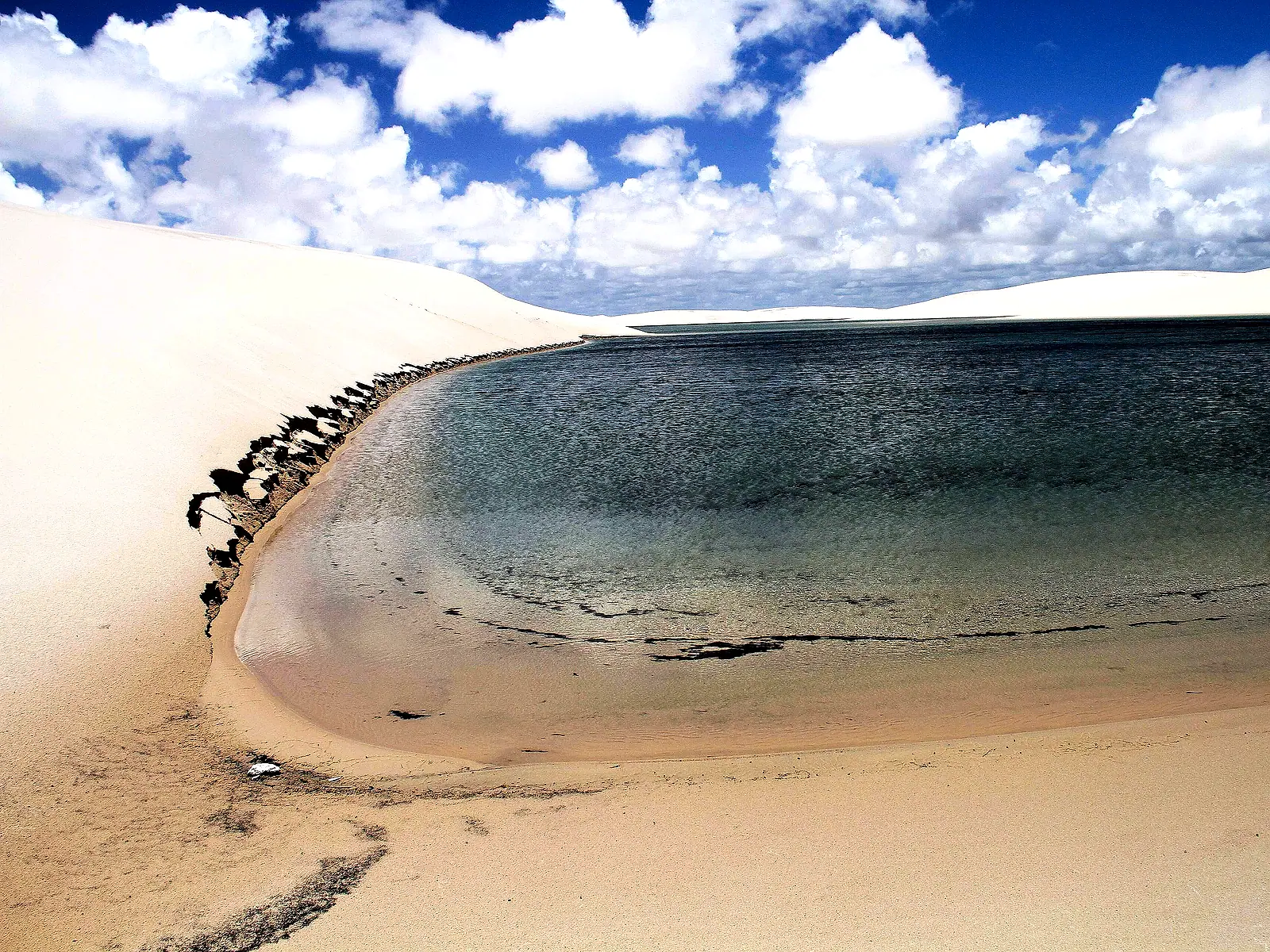 Lagoa de água transparente na curva de uma duna branca, sob céu azul com nuvens