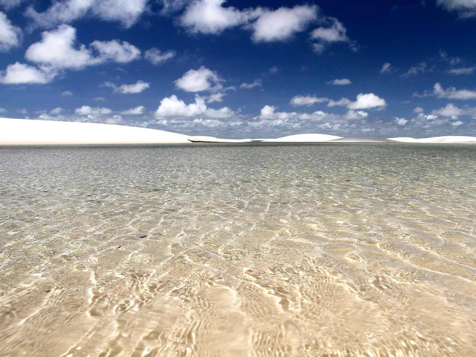 Água transparente da Lagoa Emendada sobre a areia, com dunas e céu azul ao fundo
