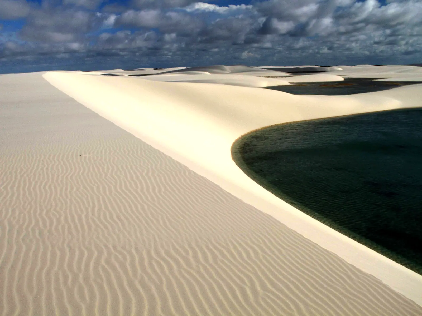 Duna de areia branca contornando uma lagoa de água escura sob céu com nuvens