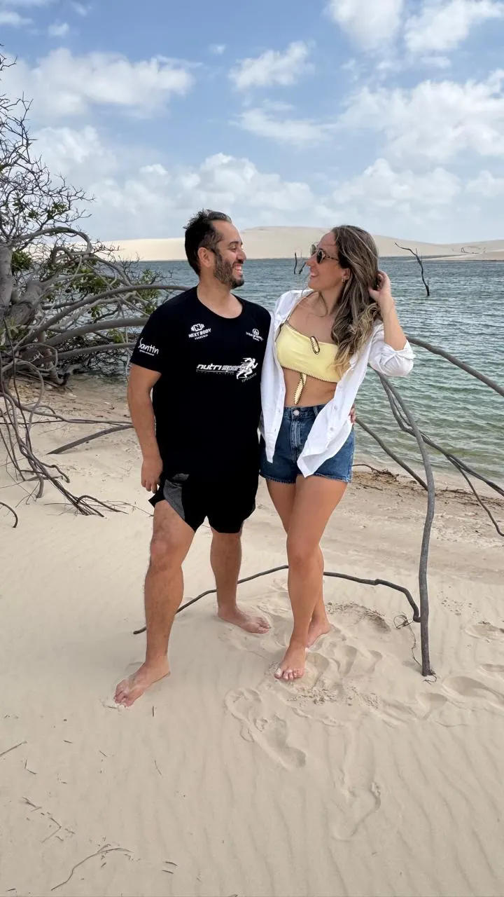 Casal na areia à beira de uma lagoa, com galhos secos e dunas ao fundo